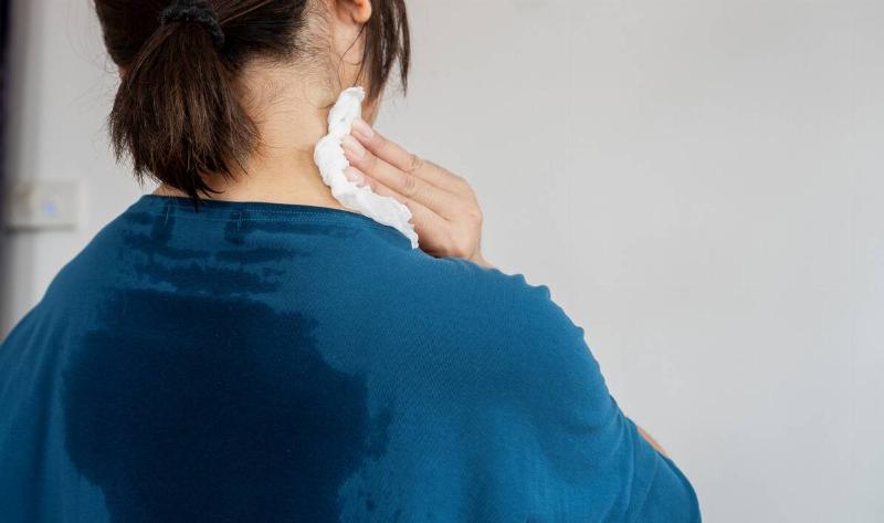 A woman visibly sweating through her shirt on her back, wiping the side of her neck with a cloth.