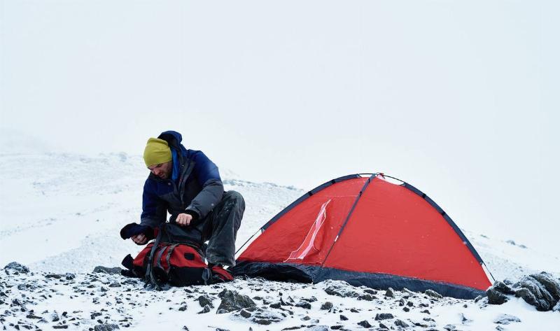 A man outside in the snow, a small tent behind him, looking through his hiking pack.