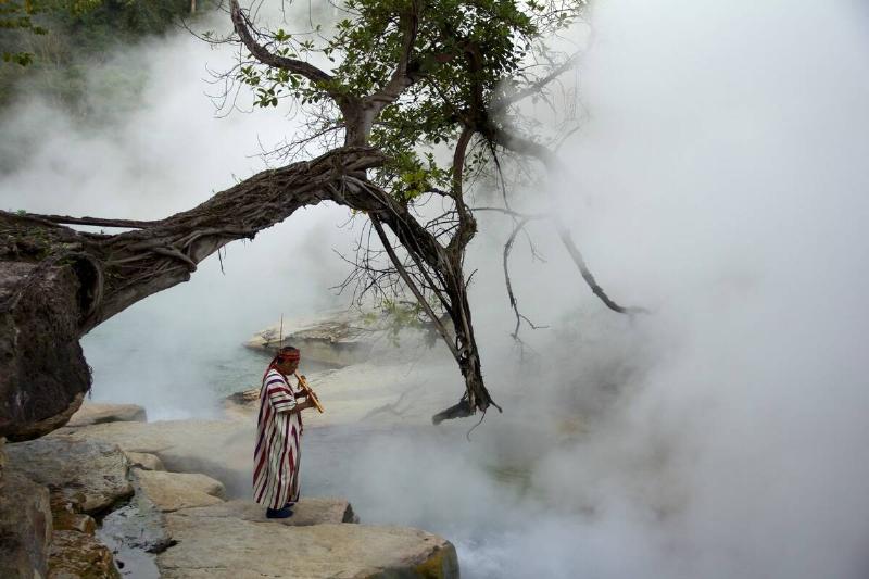 A man standing near the Boiling River, playing a wind instrument.