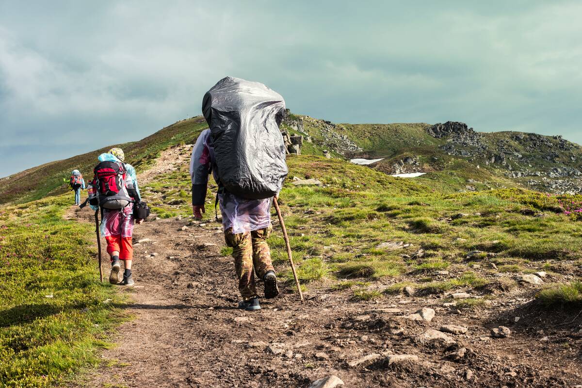 Three hikers with backpacks walking in mountain trail, the one at the back sporting a large plastic cover over their pack.