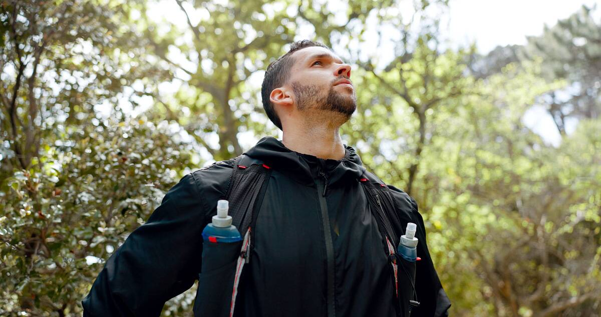 A low-angle shot of a man hiking outdoors, two water bottles fastened to his backpack straps.
