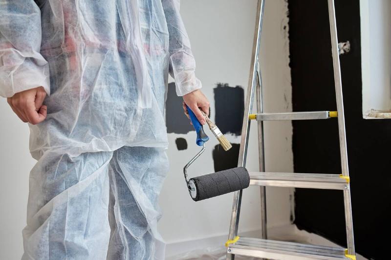 Woman wearing protective coveralls holds black paint roller near painted  wall samples and ladder in room under renovation. Painter preparing to 
paint wall. Home improvement and interior update concept