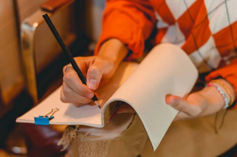 unrecognizable caucasian woman sitting writing with a black pencil on a blank notepad on her legs, copy space on the right