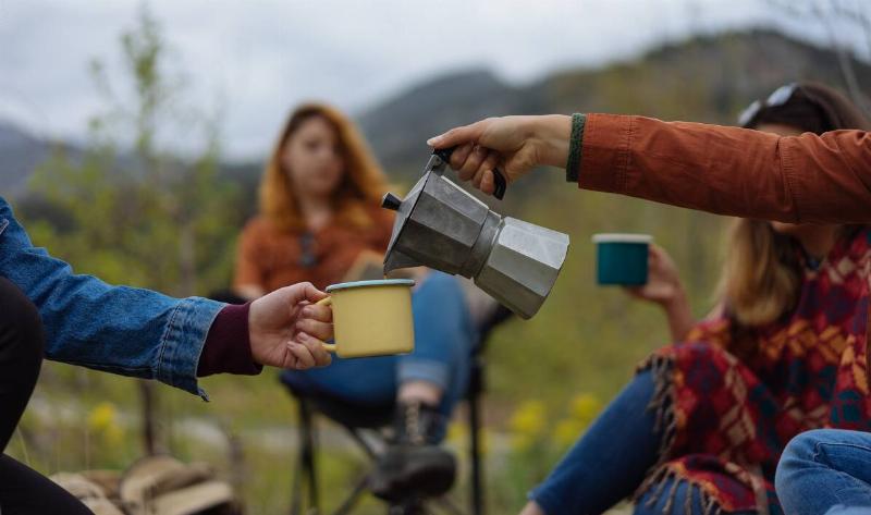 People camping, one pouring coffee from a moka pot into another's mug.