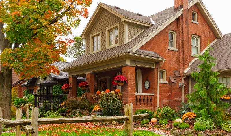 A home in the autumn, a tree shedding leaves around it, pumpkins on the porch railing.