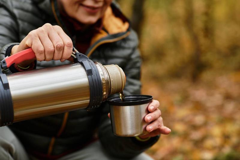 A senior woman pours a warm drink from a thermos into her cup amidst colorful autumn foliage.