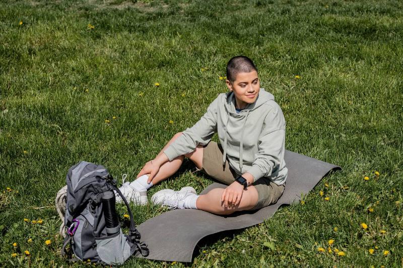 Smiling short haired female tourist sitting on fitness mat near backpack with travel equipment and looking away on grassy lawn with flowers, finding serenity in nature, summer