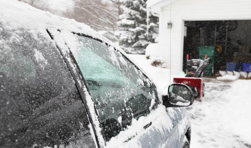 A snow-covered car in a driveway, a snowblower seen in front of it.