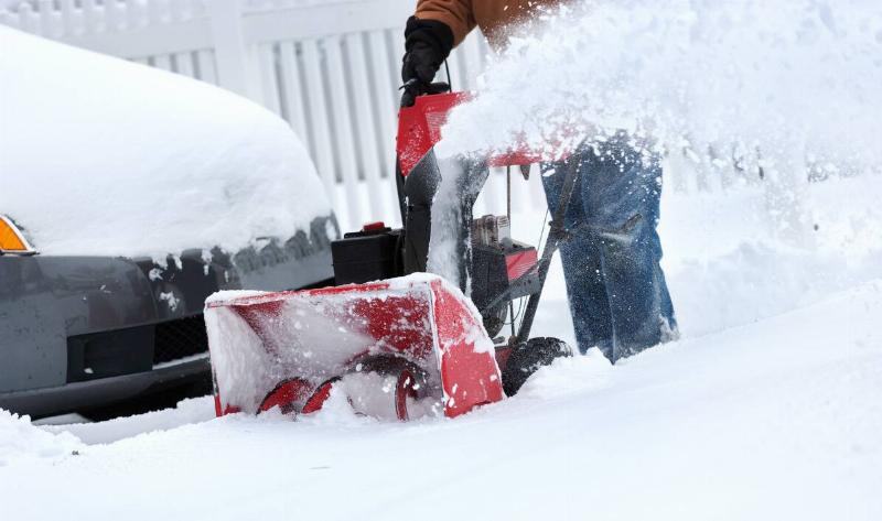 A man pushing a snowblower around a car.