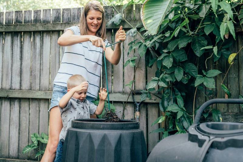 A mother turning her compost bin with her young son watching.