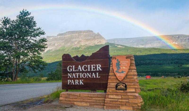 A sign for Glacier National Park, a rainbow visible in the sky behind it.