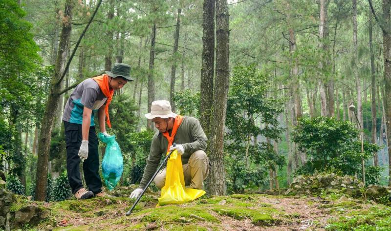 Two people volunteering by picking up garbage in the forest, a man kneeling on the ground with a younger boy standing next to him.