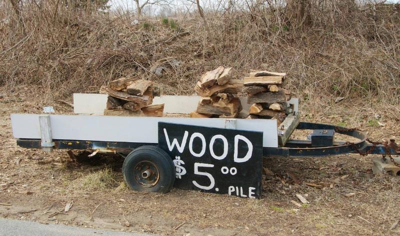 A flatbed tow trailer with stacks of wood in it. On the ground in front of the trailer is a hand-painted sign that says 