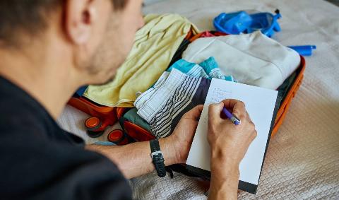 A man crossing things off a list as he packs a suitcase.