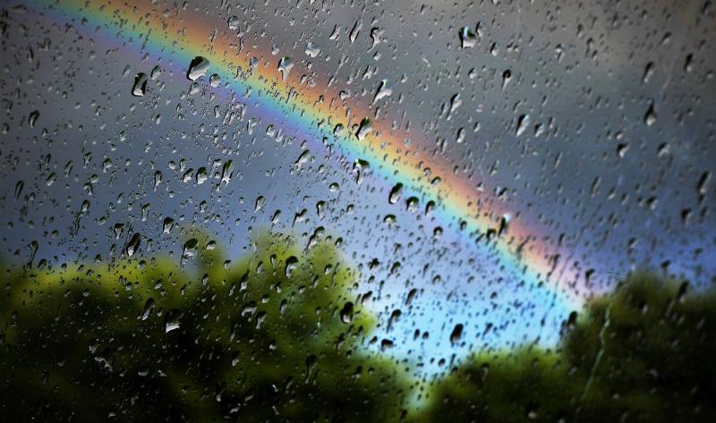 A window covered in rain drops, a rainbow visible out of focus through it.