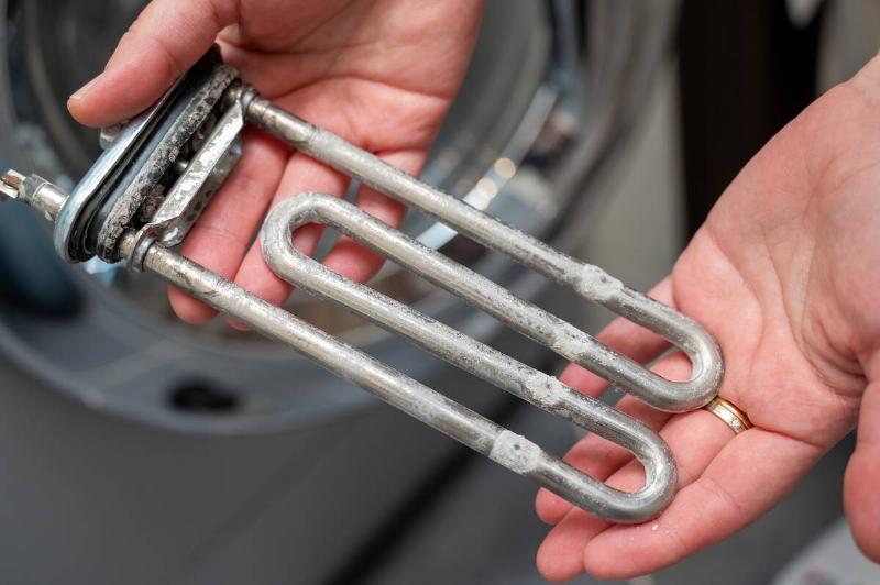 Close-up of a person holding a washing machine heater element with heavy  limescale buildup, illustrating repair, cleaning, and maintenance of 
household appliances