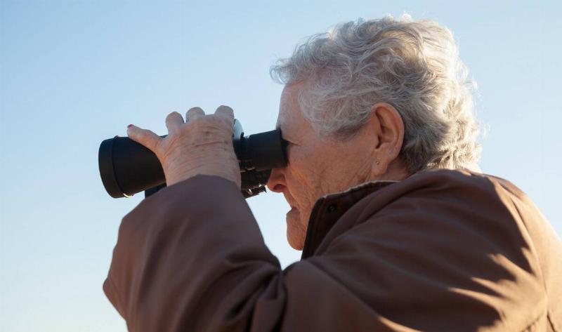 A close photo of an older person looking through a pair of binoculars.