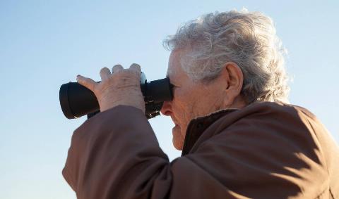 A close photo of an older person looking through a pair of binoculars.