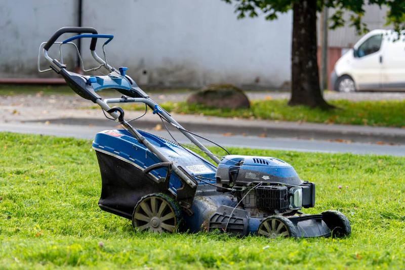 A blue lawn mower sits on lush green grass near a residential street on a sunny day.