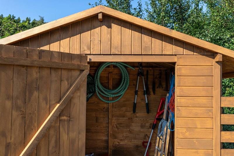 A wooden garden shed standing on a concrete foundation in a garden, visible garden tools.