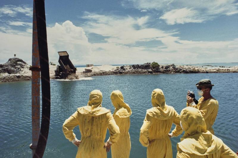 German-born American military officer Herbert E Wolff and military personnel of the 84th Engineers Battalion, 45th Support Group, USASCH, each wearing protective yellow coveralls as they watch as hardened concrete mixed with contaminated soil and attapulgite additive being dumped from a 20-ton truck into the crater formed by a nuclear bomb, on the northern tip of Runit Island, one of forty islands of the Enewetak Atoll, Marshall Islands, 26th July 1978.