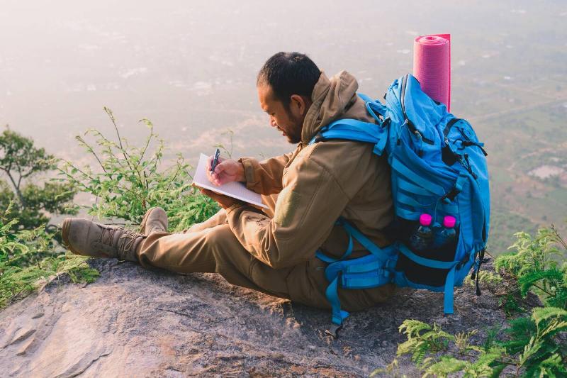A man sitting on a hill with a backpack and writing in a journal. 