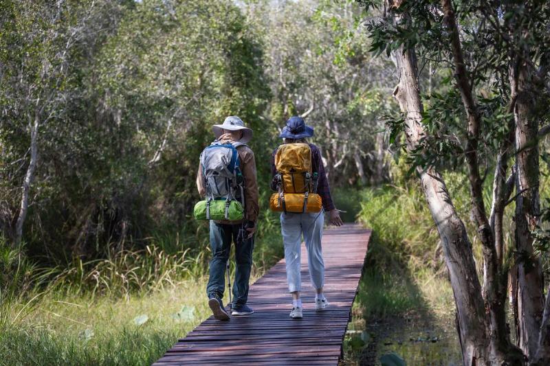Two hikers with backpacks walk along a wooden boardwalk through a lush forest.