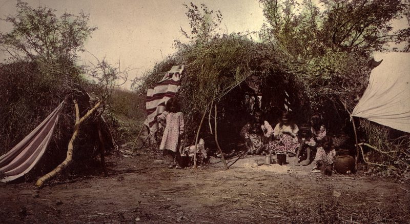View of a Native American Apache camp, Arizona, shows a Chiricahua Apache medicine man with his family inside a brush wickiup.