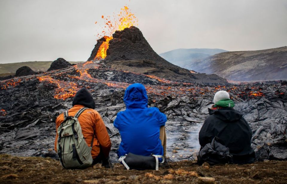 Three individuals sitting in front of the Fagradalsfjall volcano while it's erupting