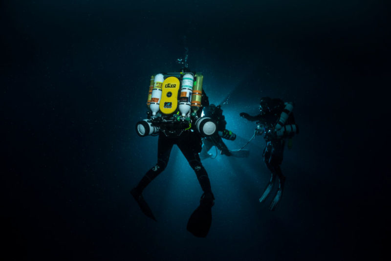 Three divers swimming in dark water, illuminated only by a powerful light