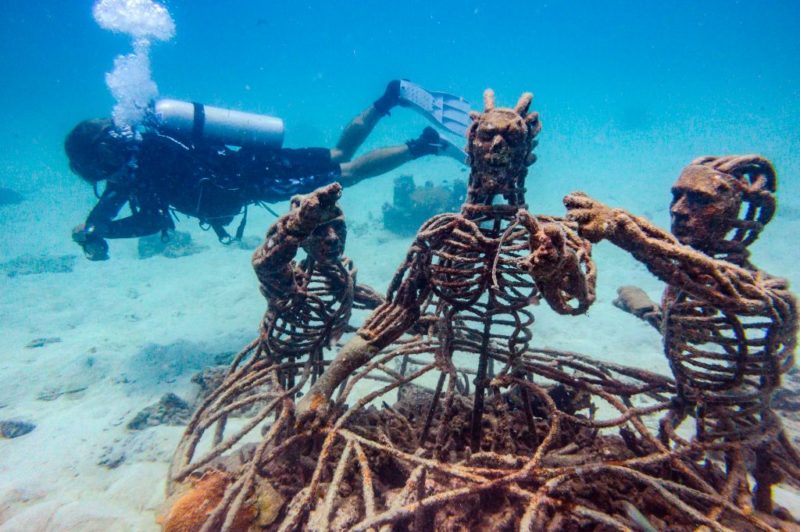 Diver swimming behind a sculpture of three skeleton-like figures