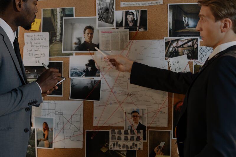 Two detectives standing in front of a cork board covered in images